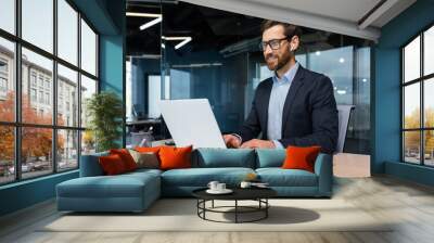 Mature businessman in headphones small earbuds talking on a video call using a laptop, boss at work at the desk in a business suit in the middle of the office smiling friendly. Wall mural