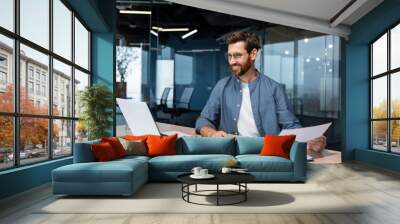 Mature businessman in shirt doing paperwork, man working with documents, contracts and bills sitting at table using laptop at work, financier accountant with beard and glasses. Wall mural