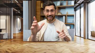 Mature teacher teaching online remotely, man with glasses and beard looking at web camera and smiling talking to colleagues and teaching, businessman at home sitting on sofa in living room. Wall mural