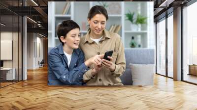 Mother and son sitting together on sofa at home in living room, family looking at phone screen watching video online and choosing shopping in online store hugging and smiling close up. Wall mural