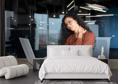 Overtired business woman working inside office with laptop, Hispanic female worker has severe neck pain, massaging muscles with hand. Wall mural