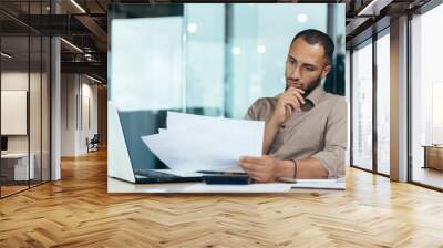 Pensive serious businessman reading financial report, hispanic businessman holding document in hands looking disappointed, working inside modern office with laptop behind paper work. Wall mural