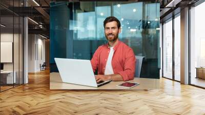 Portrait of a smiling and successful mature man in the office, a businessman with a beard and a red shirt is smiling and looking at the camera, a programmer is using a laptop at work, a creative Wall mural