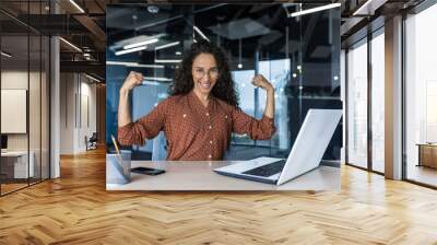 Portrait of a strong business woman female worker working inside office building, sitting a desk and using a laptop, smiling and looking at the camera holding hands up gesture superpower and success Wall mural