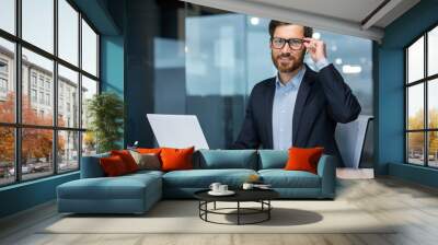 Portrait of a successful young businessman, lawyer, legal defender. Confidently looks into the camera, holds glasses, laughs, sits in the office at the table, works at the laptop. Wall mural