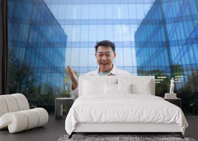 Portrait of a young Asian man standing near a glass skyscraper and holding a phone. Happy received good news, reads messages, chats on video call. Wall mural