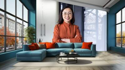 Portrait of a young successful and confident Asian female programmer, developer and IT expert. He stands in the office center with his arms crossed on his chest and looks at the camera with a smile. Wall mural