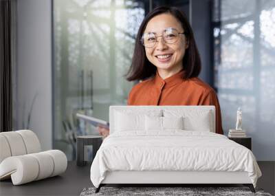 Portrait of a young successful Asian female programmer inside office, worker in glasses smiling and looking at the camera, a businesswoman holds a tablet computer in hands and uses it to test online . Wall mural