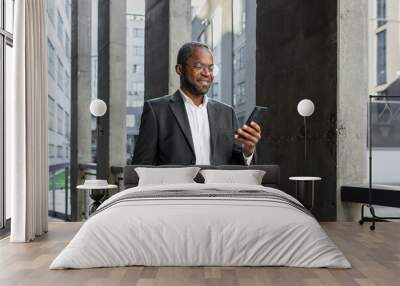 Portrait of an African American businessman, banker standing in a suit outside an office building and holding a phone. He keeps his hand in his pocket, reads news, writes a message, checks his mail. Wall mural