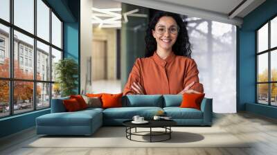 Portrait of happy and successful business woman, boss in shirt smiling and looking at camera inside office with crossed arms, Hispanic woman with curly hair in corridor Wall mural