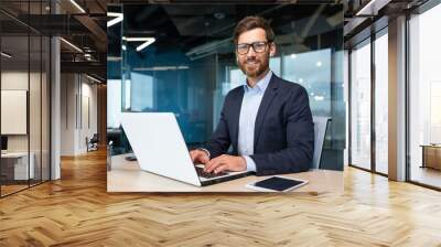 Portrait of mature boss inside office with laptop, successful and satisfied investor manager looking at camera and smiling man in glasses and business suit, investor with beard sitting on chair Wall mural