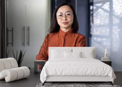 Portrait of mature experienced asian businesswoman boss, female worker looking at camera seriously, woman in glasses with crossed arms inside office building confident of successful achievement. Wall mural