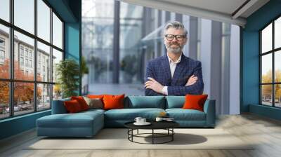 Portrait of mature gray-haired businessman, senior boss smiling and looking at camera with arms crossed, gray-haired man outside office building, successful investor and owner in business suit. Wall mural