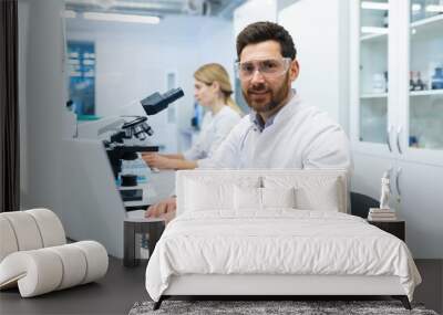 Portrait of mature successful scientist lab technician at workplace in laboratory with microscope, senior man with beard and goggles smiling and looking into camera, inventor with laptop . Wall mural