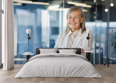 Portrait of mature successful senior woman boss inside office in business suit, businesswoman smiling and looking at camera, standing by window with crossed arms, banker finance woman. Wall mural