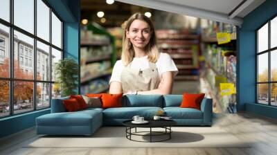 portrait of saleswoman, woman smiling and looking at camera in supermarket. Pleasant friendly female seller standing in the store between the rows . Trade business and people concept Wall mural