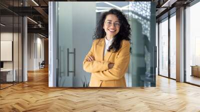 Portrait of successful business woman inside office, latin american boss in yellow suit smiling and looking at camera, mature female worker with curly hair standing with arms crossed. Wall mural