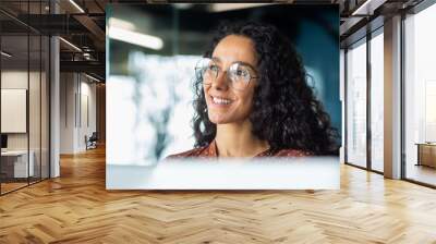 Portrait of successful businesswoman wearing glasses inside office, woman looking out window smiling and dreaming happy with achievement results, Hispanic woman at workplace with laptop. Wall mural