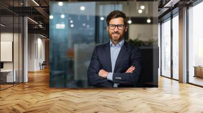 Portrait of successful mature boss, senior businessman in glasses and business suit looking at camera and smiling, man with crossed arms working inside modern office building. Wall mural