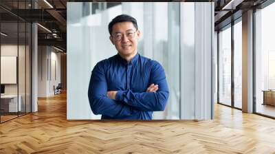 Portrait of successful mature boss, senior businessman in glasses Asian looking at camera and smiling, man with crossed arms working inside modern office building. Wall mural