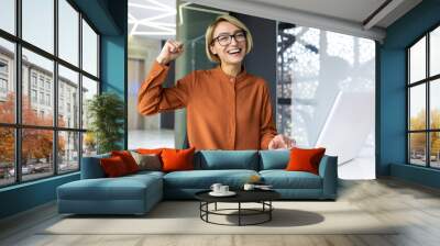 Portrait of successful satisfied woman inside office at work, business woman working on laptop, joyfully looking at camera, contentedly holding hands up, celebrating achievement of results. Wall mural