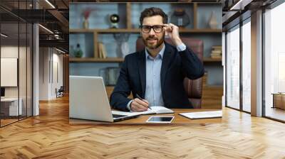 Portrait of successful smiling man in office, mature businessman looking at camera cheerfully, senior boss in glasses and beard working inside office with laptop and documents, signing contract. Wall mural