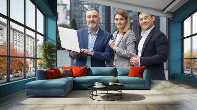 Portrait of three diverse business colleagues, business group outside office building, asian man and woman looking at camera and smiling, dream team with laptop Wall mural