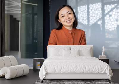 Portrait of young Asian successful business woman, female worker at workplace inside office smiling and looking at camera, video call online consultation listening to interlocutor with smile. Wall mural