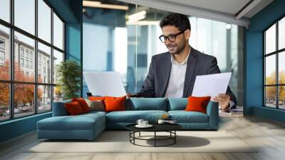 Portrait of young financier businessman doing paperwork, man in business suit checking report, contract, account, employee inside office working with laptop at workplace, smiling satisfied. Wall mural