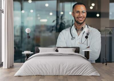 Portrait of young happy and smiling doctor, man in medical coat and stethoscope smiling and looking at camera, Arab doctor with crossed arms working inside modern clinic office. Wall mural