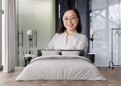 Portrait of young mature asian businesswoman entrepreneur, woman with crossed arms smiling and looking at camera, working inside office at workplace, standing near window in light clothes. Wall mural