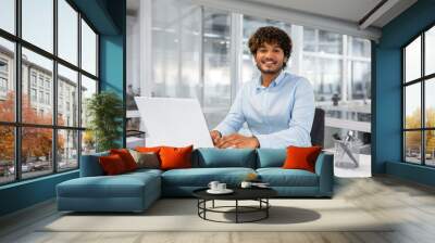 Portrait of young successful contemporary businessman inside office, man at workplace smiling and looking at camera, young hispanic businessman sitting at desk using laptop. Wall mural