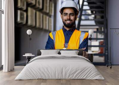 Portrait of young successful engineer worker, man smiling and looking at camera, wearing hard hat and vest, working in factory plant. Wall mural