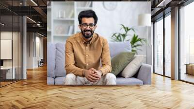 Portrait of young successful hispanic man at home on couch in living room, man smiling and looking at camera Wall mural