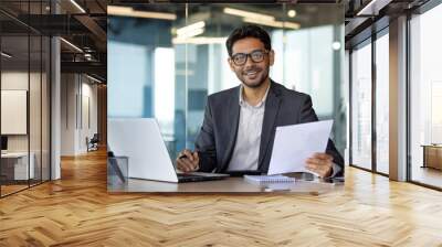 Portrait of young successful Indian businessman financier, man smiling and looking at camera, working sitting at laptop, holding reports, papers and contracts, satisfied investor entrepreneur. Wall mural