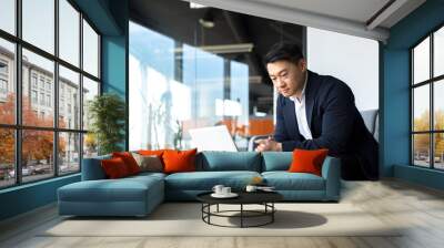 Portrait thoughtful Asian businessman working on a laptop computer at a modern office desk. Confident Focused pensive man in formal suit indoors. thinking of inspiration solving a problem. Startup Wall mural