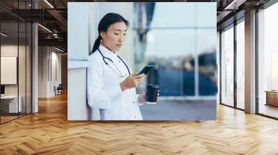 Portrait young Asian doctor woman or paramedic stands exhausted on a break near the wall of a hospital clinic with a using mobile phone in her hands. intern woman uses smartphone outside Wall mural