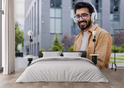 Portrait young Indian man with headphones and tablet watching online video sitting on bench near office building businessman smiling and looking at camera, portrait of satisfied online stream viewer Wall mural