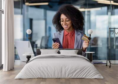 Satisfied african american female keeping debit card and cellular phone in hands by desk with computer and notebook. Pleased lady entering bank data in application for completing cashless payment Wall mural