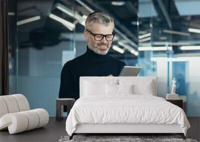 Senior and successful smiling gray-haired businessman in modern office holding tablet computer, man standing near window reading news, happy boss in glasses and beard typing message Wall mural