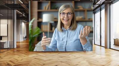 Senior woman with glasses engaging in a video call while looking and talking to the camera. She seated in a cozy living room with shelves and plants in the background, smiling warmly and gesturing. Wall mural