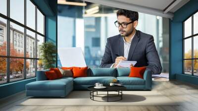 Serious and concentrated thinking businessman watching webinar online course, man upgrading at workplace inside office, hispanic man taking notes and writing data in notebook. Wall mural