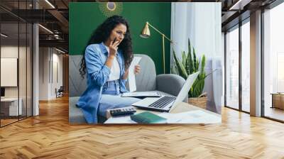 Serious and thoughtful businesswoman working at home with documents, hispanic woman in casual clothes using laptop at work sitting on sofa in living room with bills and contracts. Wall mural