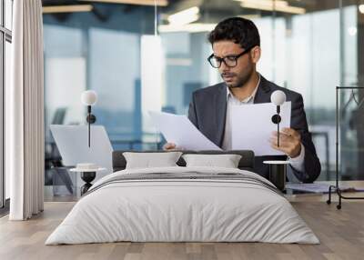 Serious concentrated businessman inside office behind paper work, financier reading and checking reports holding in hands, man thinking about technical solutions at workplace. Wall mural