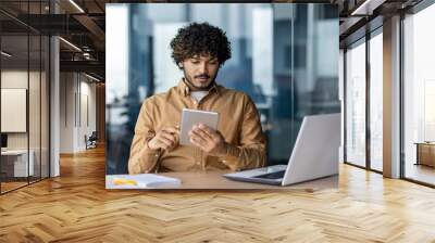 Serious focused businessman working with tablet computer sitting at desk inside office, hispanic thinking with laptop, man solving technical problem, using online application. Wall mural