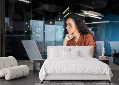 Serious thinking Latin American woman working inside office, businesswoman pondering complex decisions using laptop at work. Wall mural