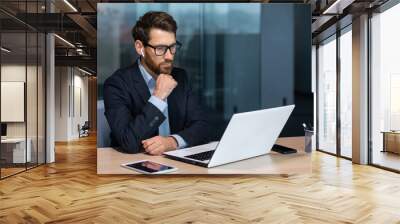 Serious thinking mature businessman working inside modern office building, senior boss in business suit and glasses working sitting at laptop, man with beard thinking about future strategy plan. Wall mural