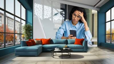 Shocked and upset business woman working inside office at workplace, depressed hispanic woman reading bad news using app on smartphone, holding phone. Wall mural