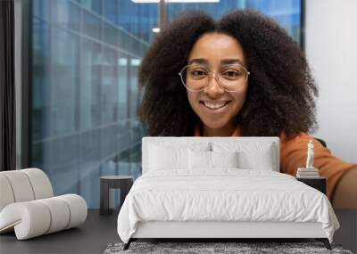 Smiling woman with curly hair taking a selfie while wearing glasses in brightly lit modern office building Wall mural