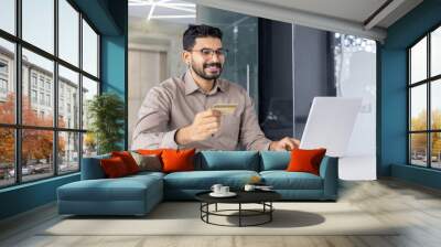 Smiling young businessman using laptop and credit card for online shopping. He is sitting in a modern office, enjoying secure digital transactions. Wall mural